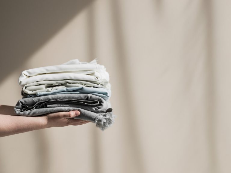 Hand holding a stack of folded CIRCLE biodegradable textiles in soft neutral and blue tones against a beige curtain